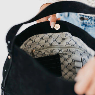 Close-up of a black suede shoulder bag being held open by hands, showing a patterned beige and black interior lining with a zipper pocket.