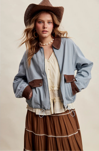 A woman wearing a light blue denim jacket with brown faux-leather trim, a cream blouse, a brown pleated skirt, and a brown cowboy hat, posed against a solid beige studio background.