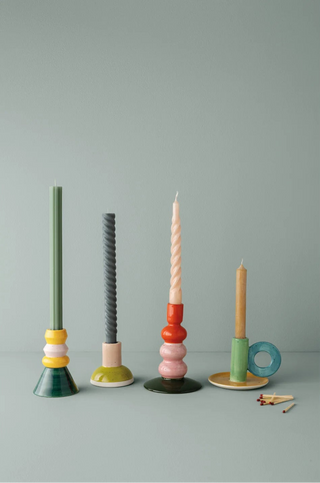 Five colorful wooden candlesticks in different designs are lined up on a light gray surface against a muted gray-green background. The set includes candles in green, gray, cream, pink, orange, and natural wood tones, with some styled with small matches.
