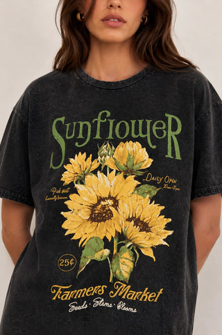 A woman wearing a dark washed graphic t-shirt featuring a large yellow sunflower print with green and gold vintage-style text reading 'Sunflower' and 'Farmers Market'.