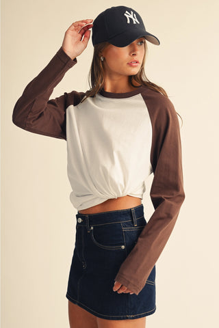 A woman modeling a long-sleeve raglan crop top with a white body and brown sleeves, paired with a dark blue denim mini skirt and a black New York Yankees baseball cap, against a solid beige background.