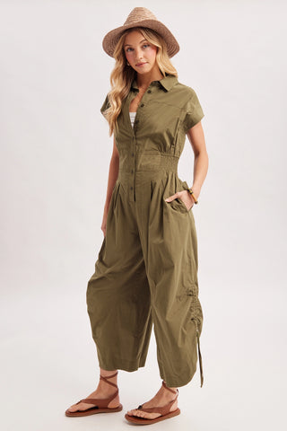 A woman modeling an olive green short-sleeve jumpsuit with a button-up front, side pockets, and gathered legs, styled with a straw hat and brown sandals against a light gray studio background.