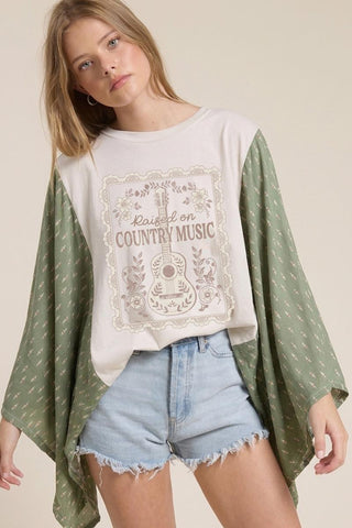 Woman wearing a white t-shirt with 'Raised on Country Music' design, green kimono-style top, and denim shorts.
