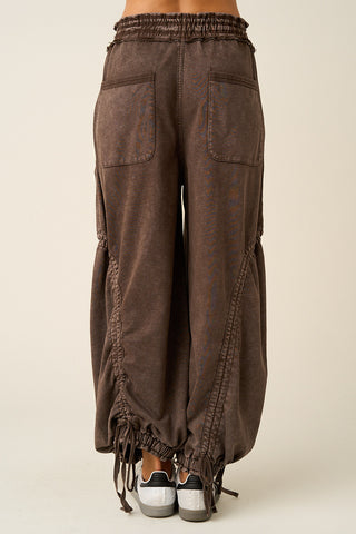 A person shown from the back wearing loose, wide-leg brown pants with an elastic waistband, large patch pockets, gathered side seams, and drawstring ties at the ankles, paired with black and white sneakers against a plain light beige studio background.