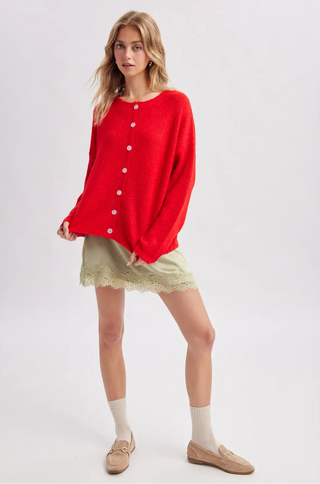 A woman modeling a bright red knit cardigan sweater with white buttons, paired with a beige lace-trimmed mini skirt, white socks, and tan loafers, against a light gray studio background.
