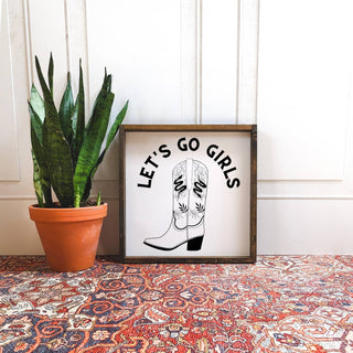 Framed print with 'Let's Go Girls' text and cowboy boots next to a potted plant on a patterned surface.