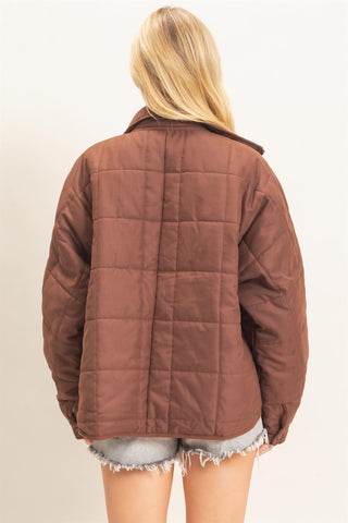A woman viewed from the back wearing a brown quilted jacket and light gray frayed denim shorts against a plain off-white background.