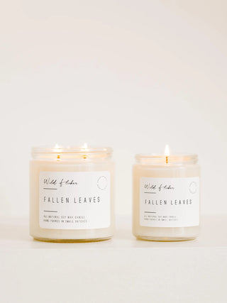 Two lit soy candles in clear glass jars with white labels reading 'Wild of Eden' and 'FALLEN LEAVES', photographed on a clean white background.