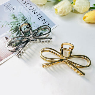 Two metal bow clips, one silver and one gold in color, placed on a surface with a floral patterned paper and white flowers in the background.
