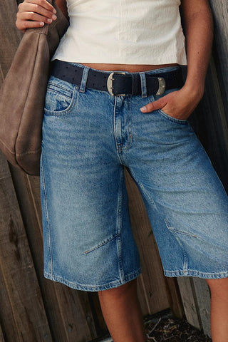 Person wearing blue denim shorts with a belt, standing against a wooden background.