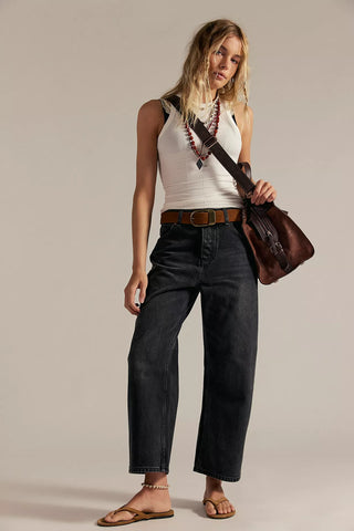 Woman wearing a white tank top, black jeans, and carrying a brown leather bag on a plain background