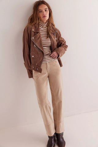 Woman wearing a brown jacket, striped shirt, beige pants, and brown shoes against a white background
