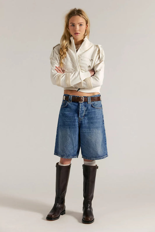 Woman wearing a white jacket, blue denim shorts, and black boots on a plain background