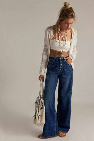 Woman wearing a white top, cardigan, and blue jeans holding a beige handbag against a plain background