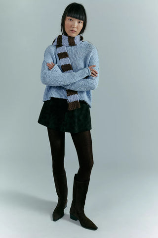 Person wearing a blue sweater, green skirt, black tights, and brown knee-high boots with a striped scarf.