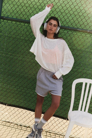Person wearing a white long-sleeve shirt and gray shorts standing next to a white chair against a green chain-link fence.