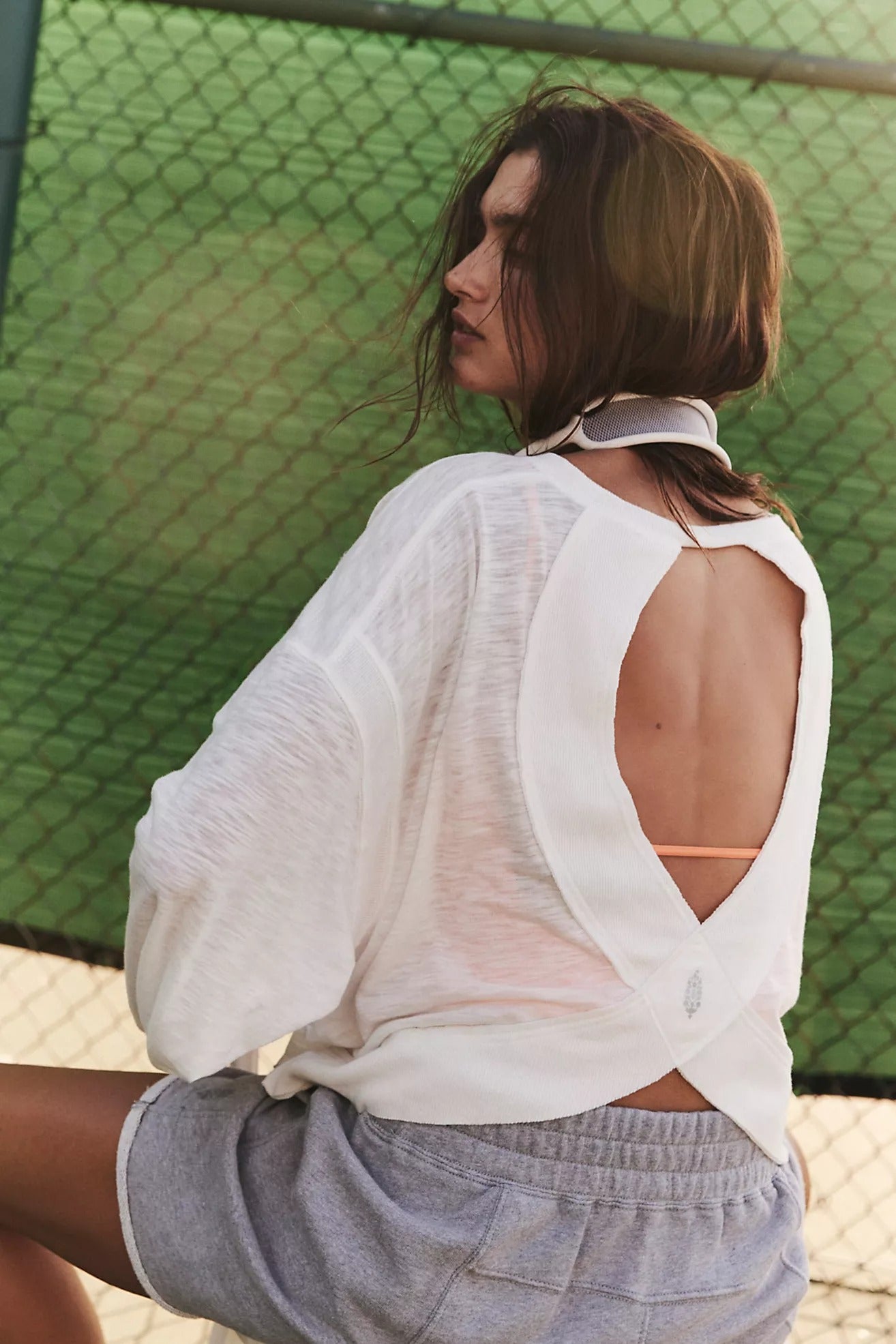 Person wearing a white open-back top and gray shorts against a green chain-link fence.