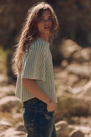 Woman wearing a light green knitted sweater and dark jeans standing in a natural setting with blurred background