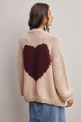 Person wearing a beige knit cardigan with a dark red heart design on a plain background