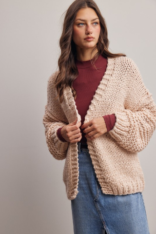 Woman wearing a beige knitted cardigan over a burgundy top and blue jeans on a plain background