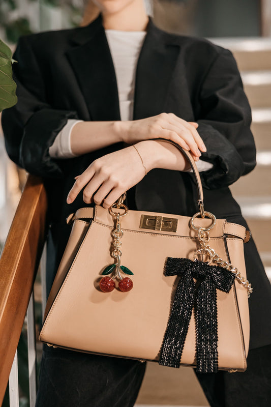 Beige handbag with decorative bow and cherries held by a person in a black coat.