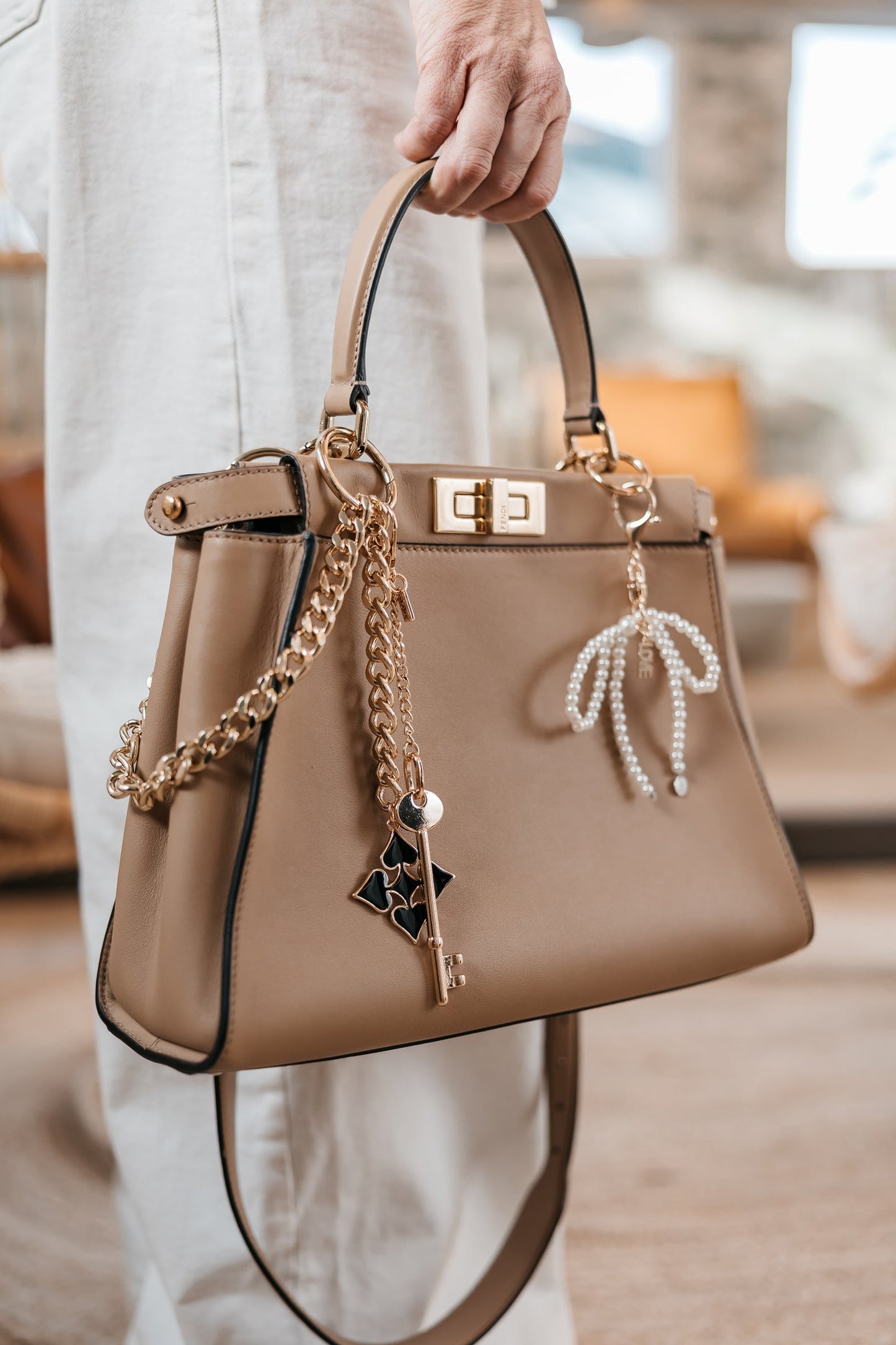 Beige handbag with gold chain and decorative elements held by a person indoors.
