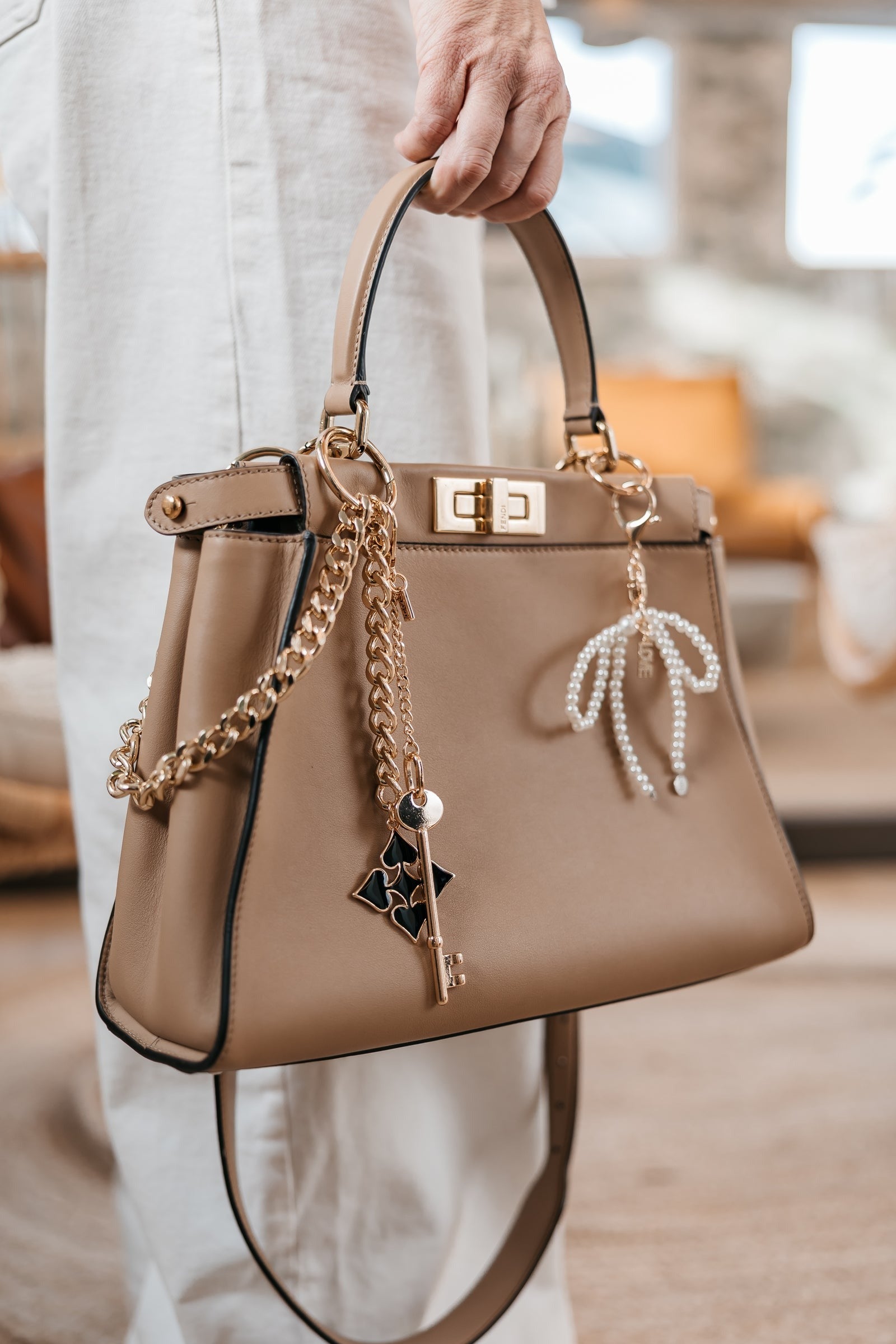 Beige handbag with gold chain and decorative elements held by a person indoors.