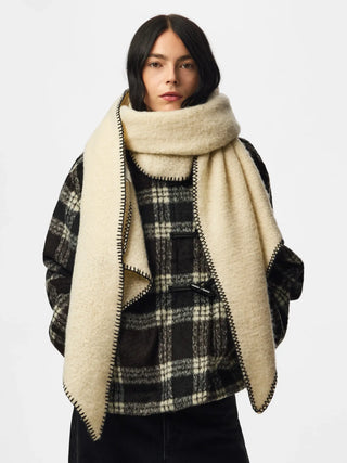 A model wearing a thick, cream-colored fuzzy scarf with black and beige zigzag trim, styled over a black and beige plaid jacket against a light gray background.