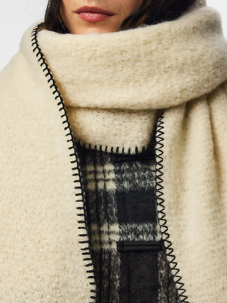 Close-up of a person wearing a thick cream-colored knit wrap or shawl with black zigzag stitching, layered over a black and beige plaid garment.