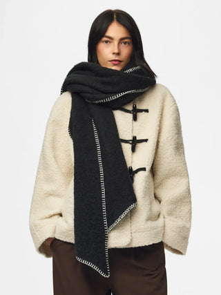A model wearing a cream sherpa jacket with toggle closures, brown trousers, and a black textured scarf with white contrast stitching and toggle-style details.