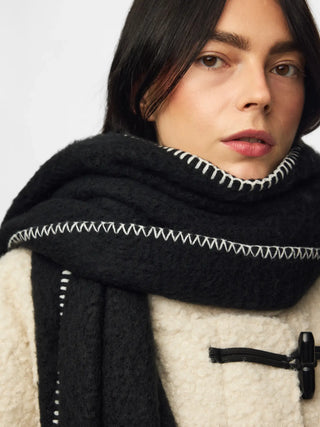 A close-up of a model wearing a thick black knit scarf with white zigzag stitching, paired with a cream-colored textured coat.