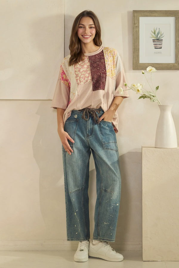 Woman wearing a patterned top and jeans standing in a room with a vase and plant in the background.