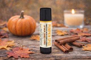 Pumpkin spice lip balm with a pumpkin, cinnamon sticks, and candle on a wooden surface.