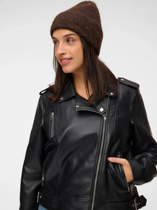 A woman wearing a brown knit beanie and a black faux-leather moto jacket, photographed against a plain light gray studio background.