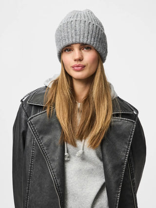 A woman wearing a light gray ribbed knit beanie, a gray hoodie, and a black leather jacket, photographed against a plain light gray studio background.