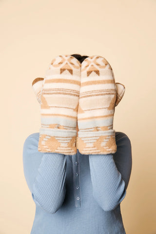 A person wearing a light blue ribbed long-sleeve top holds a pair of knit winter mittens in front of their face. The mittens have a cream, beige, and light blue Southwestern-style pattern with brown accents, set against a solid beige background.