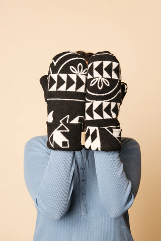 A person wearing a light blue ribbed long-sleeve top holds a pair of black knit mittens with white geometric and animal motifs in front of their face against a solid beige background.