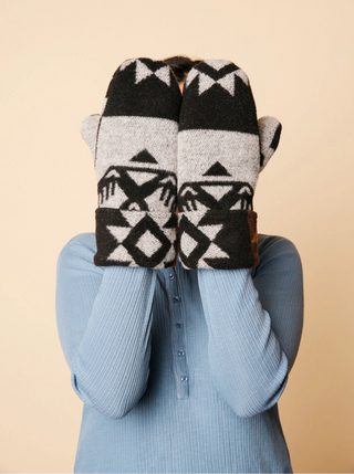 A person wearing a light blue ribbed long-sleeve top holds up a pair of knit fingerless mittens with a black, white, and gray geometric pattern over their face, against a solid beige background.