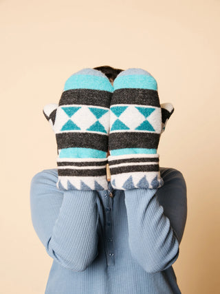 A person wearing a light blue ribbed cardigan holds up a pair of knit mittens in front of their face. The mittens feature a Southwestern-style pattern with turquoise, white, and dark gray geometric stripes and triangles, set against a solid beige background.