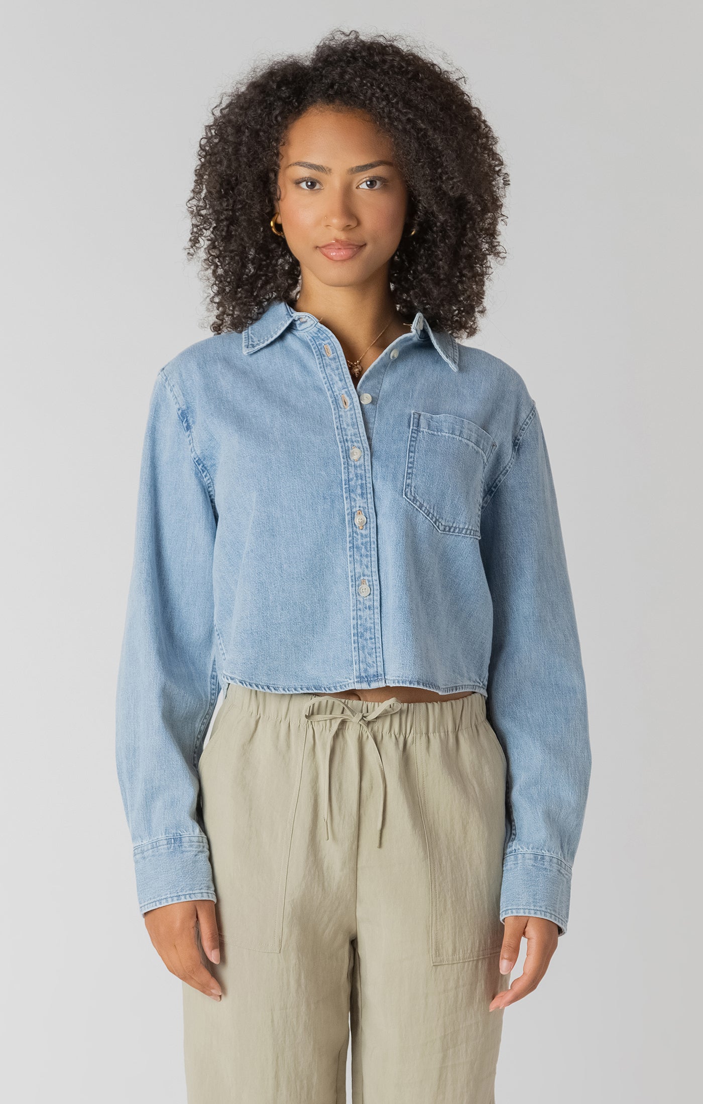 Woman wearing a light blue denim shirt and beige pants against a plain background