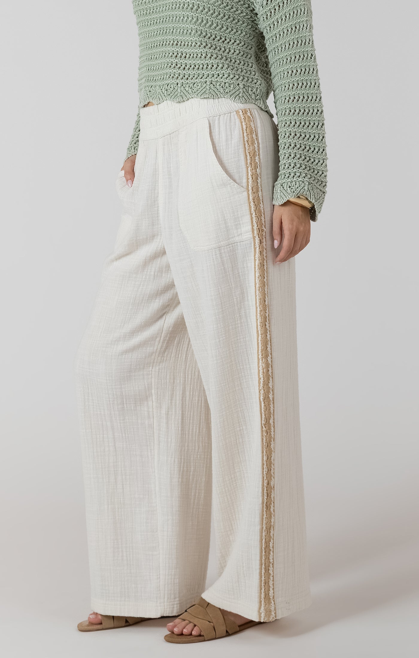 Person wearing a green knitted sweater and white pants with a gold stripe on a plain background