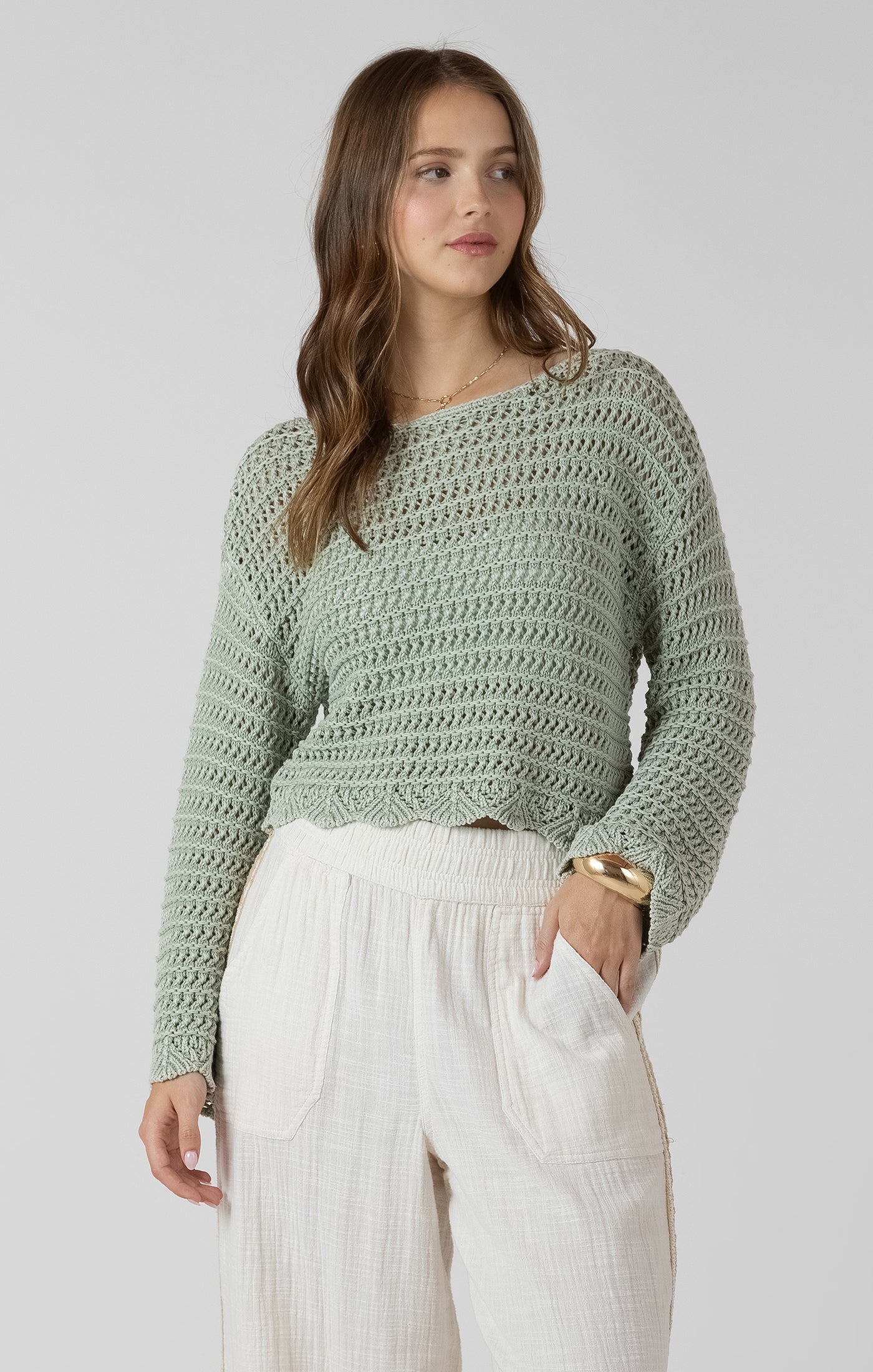 Woman wearing a green crochet sweater and white pants on a plain background