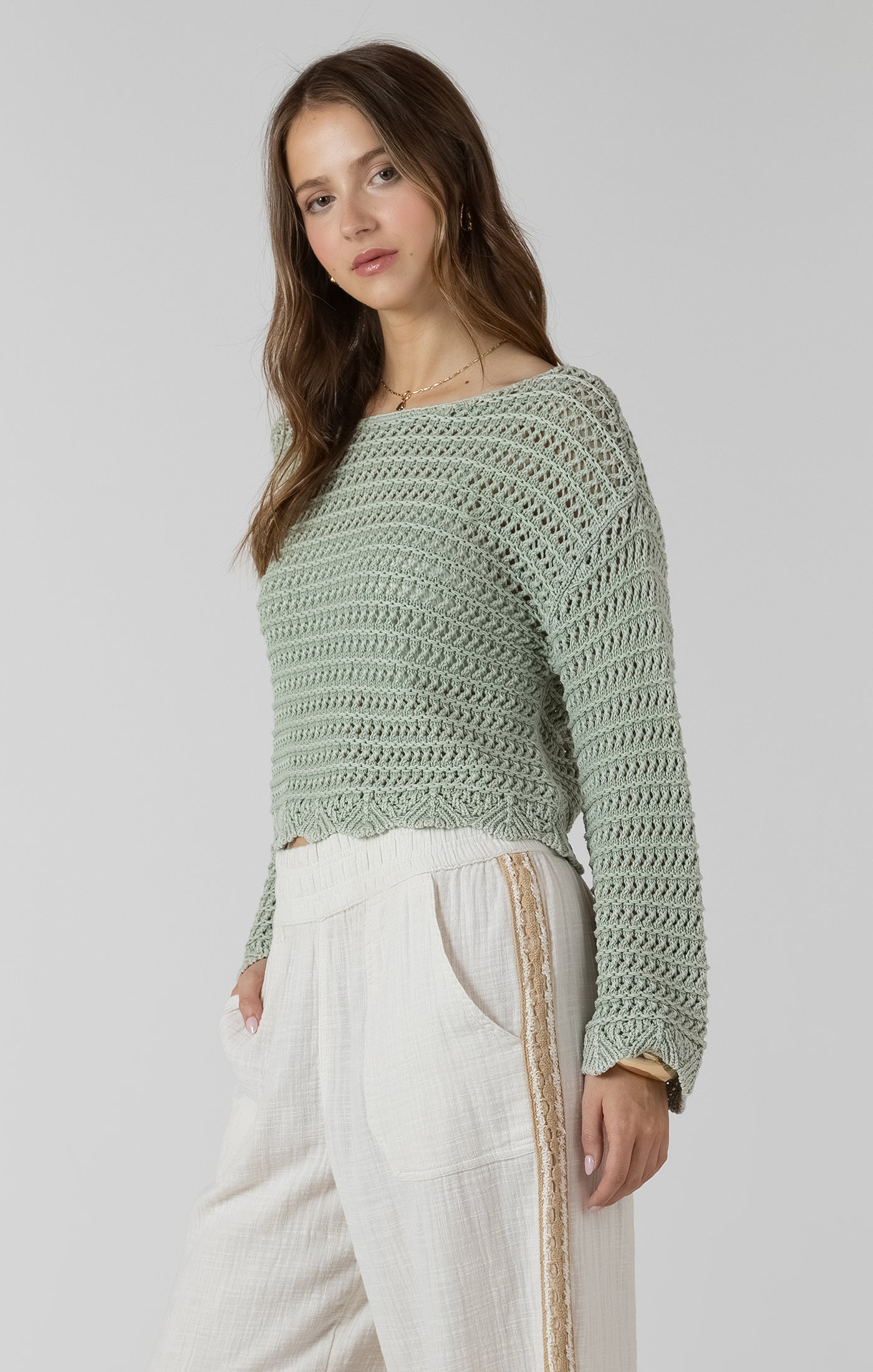 Woman wearing a green crochet sweater and white pants on a plain background