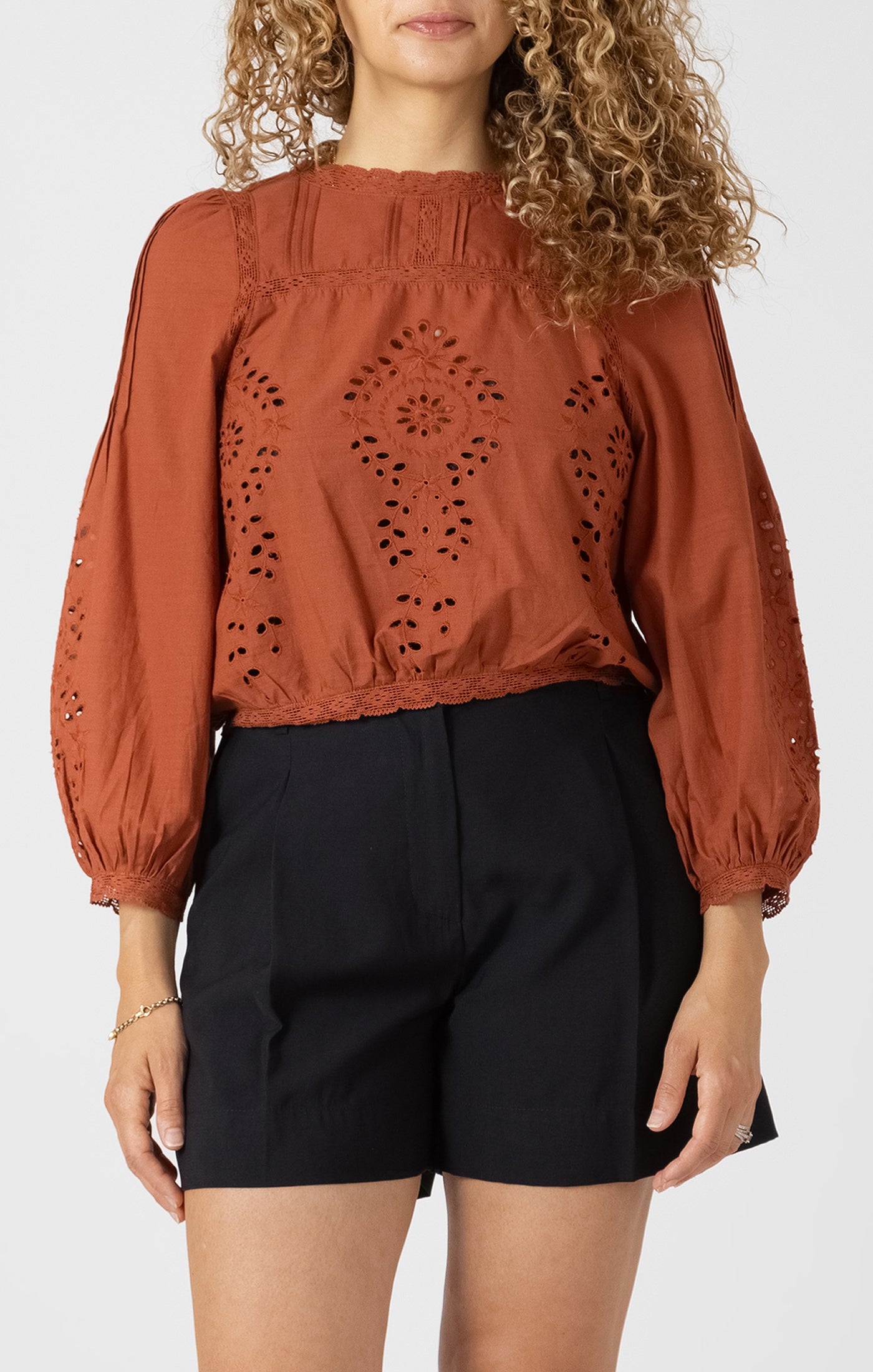 Woman wearing a rust-colored blouse with intricate cut-out design and black shorts on a white background