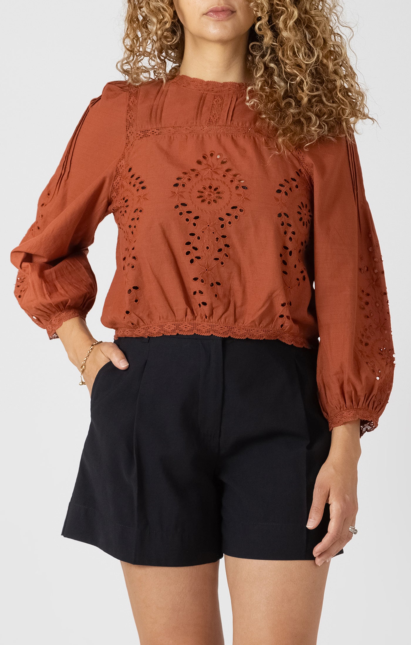 Woman wearing a rust-colored blouse with intricate cut-out design and black shorts on a white background