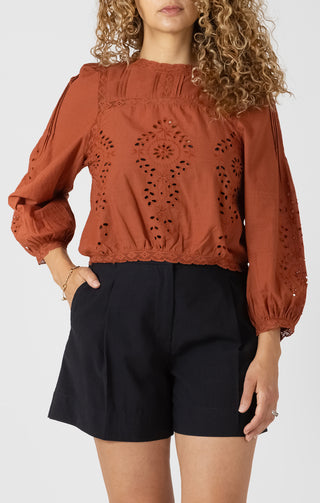 Woman wearing a rust-colored blouse with intricate cut-out design and black shorts on a white background