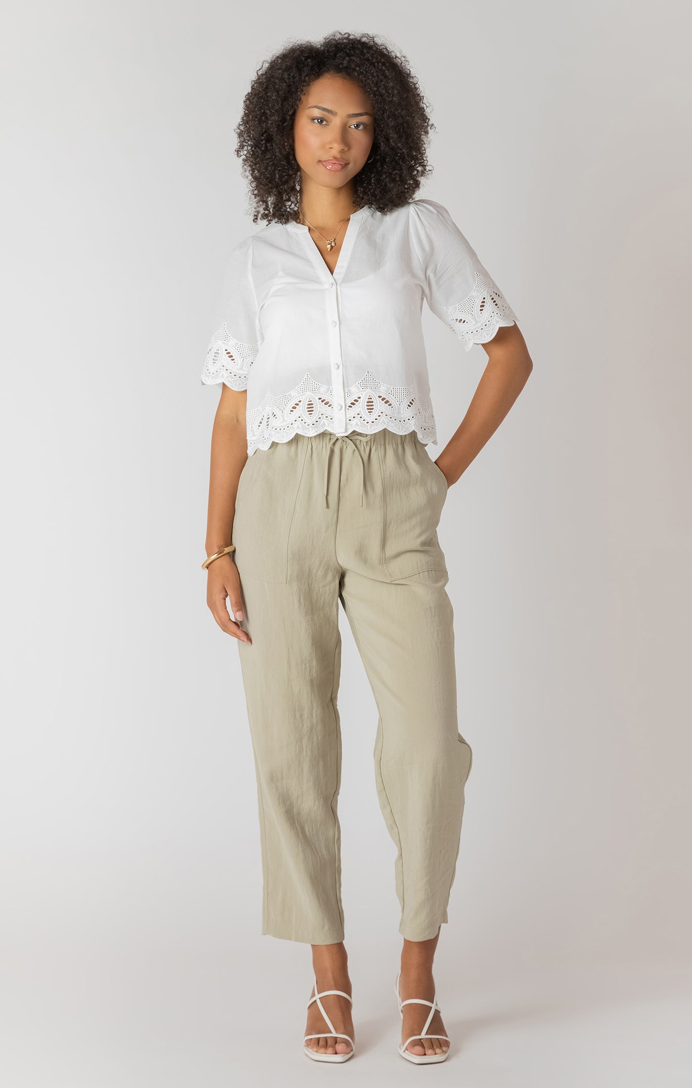 Woman wearing a white blouse with lace details and beige pants on a plain background