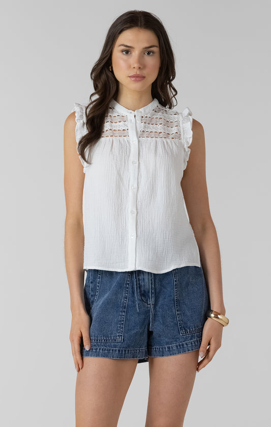 Woman wearing a white sleeveless top with lace details and denim shorts on a plain background