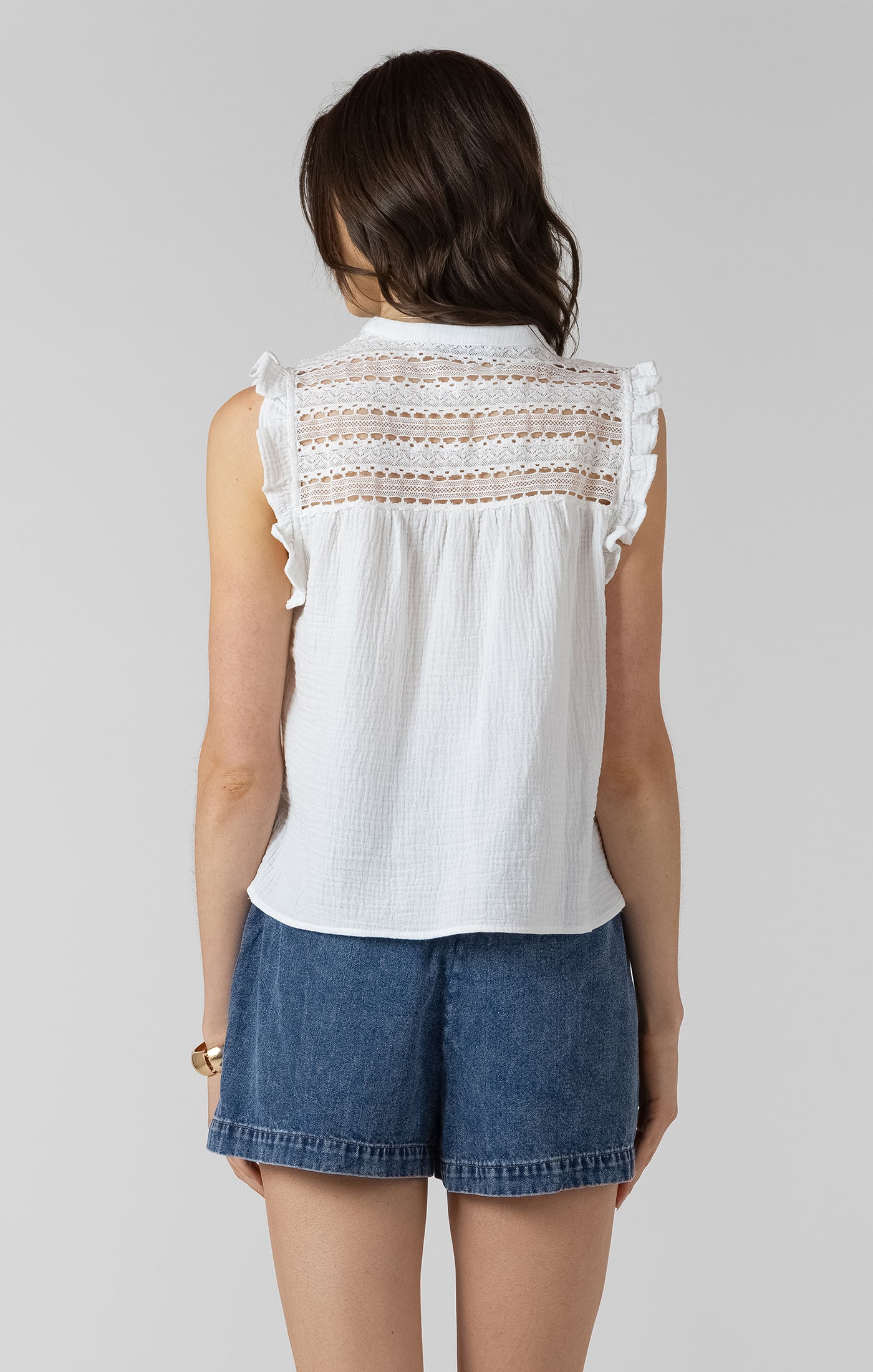 Woman wearing a white sleeveless top with lace details and blue shorts on a plain background