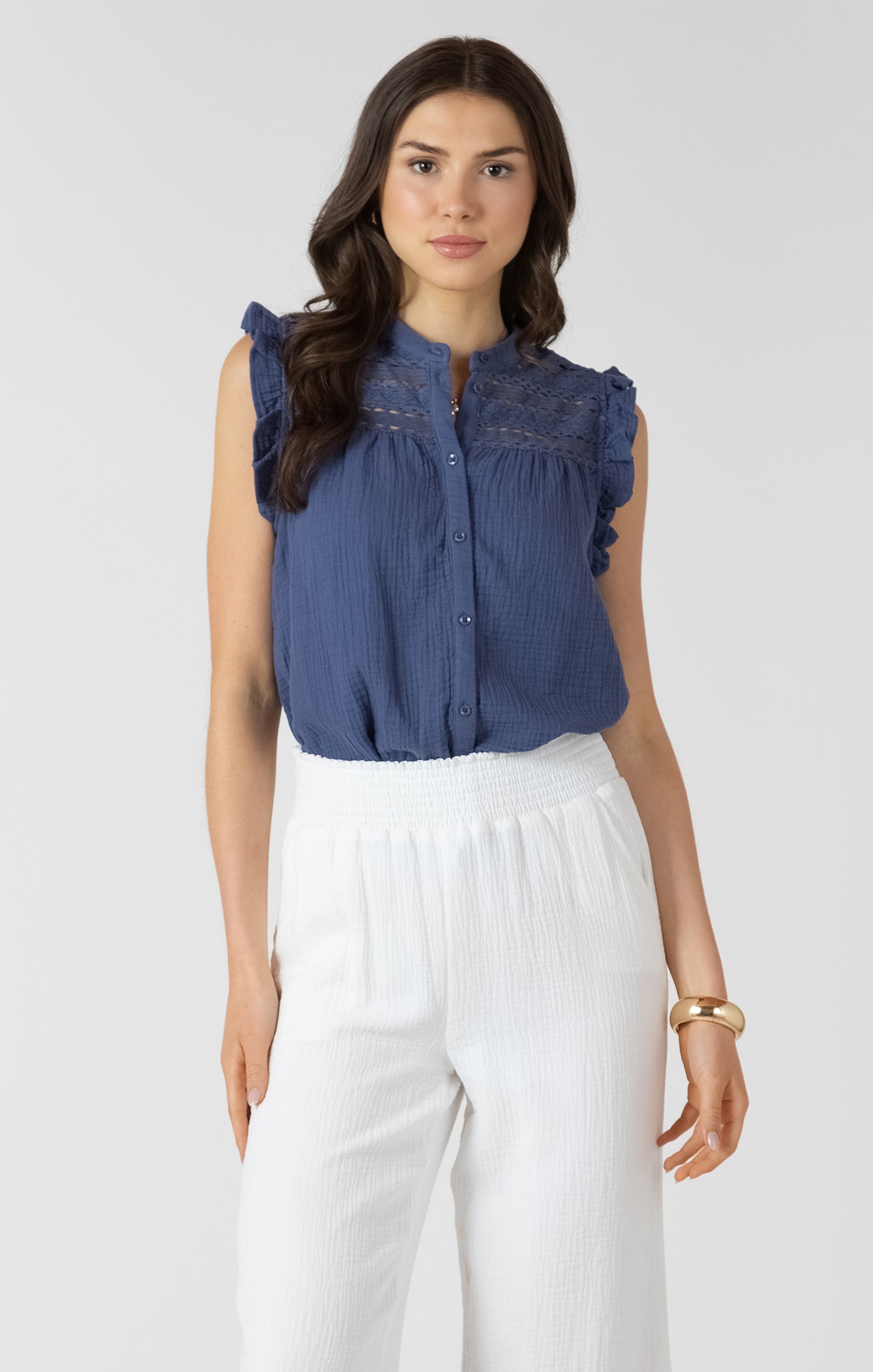 Woman wearing a blue sleeveless blouse with lace details and white pants on a white background